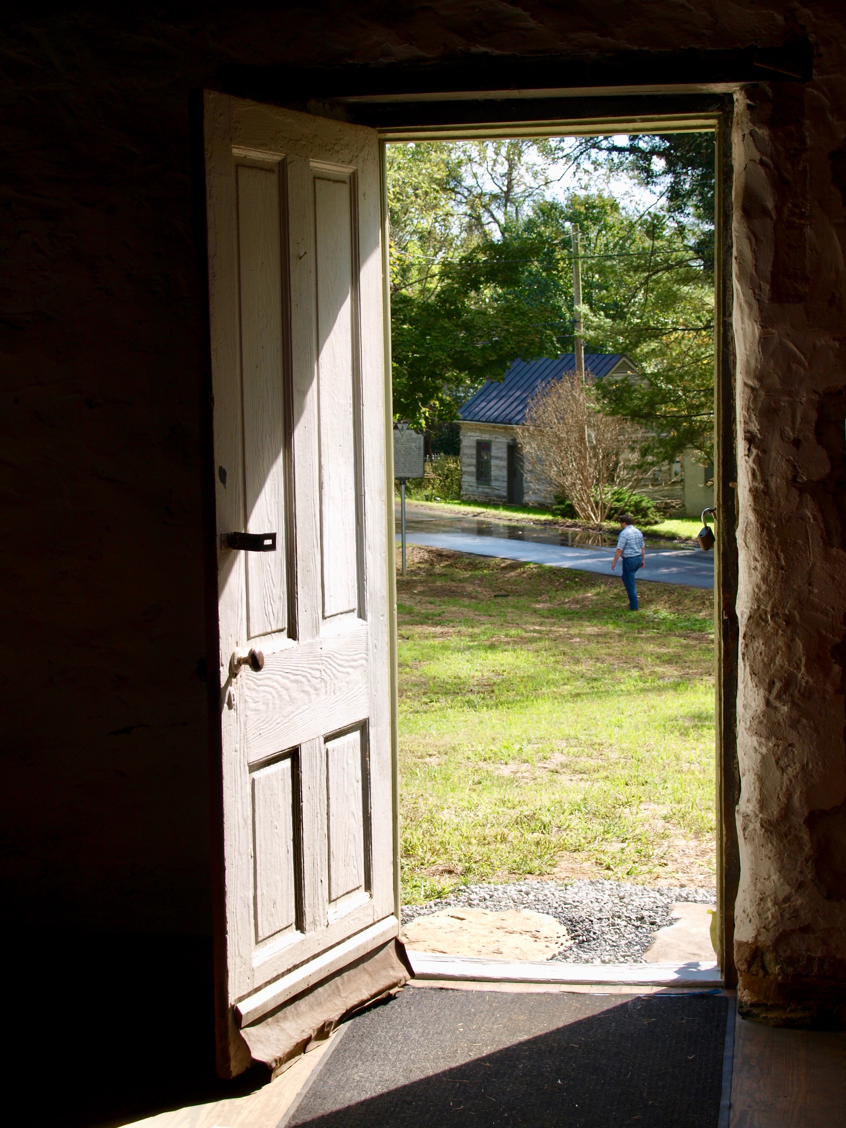 Na imagem: Porta de madeira aberta, ao fundo temos uma grama verde e um caminho indo até a porta.