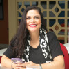 Cristina Sabbag - mulher parda, sentada sorrindo de blusa preta, cabelos escuro preso lenço laranja
